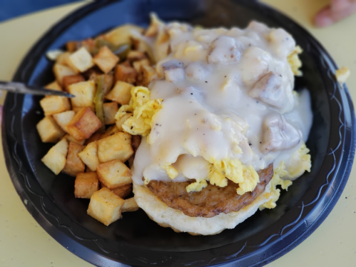 A plate with a biscuit topped with sausage, scrambled eggs, and gravy, alongside crispy potato cubes.