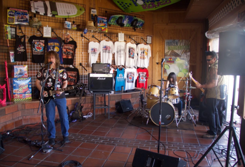 A band performs in a colorful venue with surf-themed decor, featuring guitars, drums, and merchandise on display.