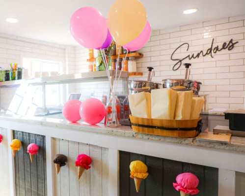 A colorful ice cream shop counter with balloons and ice cream cone decorations. The sign reads "Sundaes."