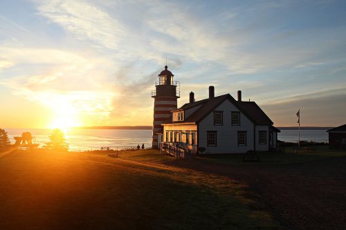 A striped lighthouse beside a white house at sunset, with a calm sea and silhouettes of people in the background.
