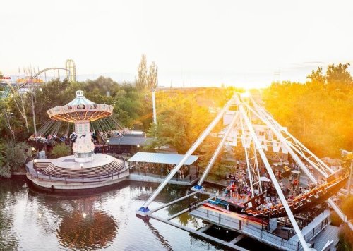 A vibrant amusement park scene at sunset, featuring a swing ride and a ferris wheel near a serene water body.