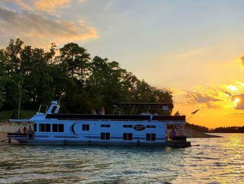 A houseboat floats on calm water at sunset, surrounded by trees and a colorful sky. People enjoy the view on deck.