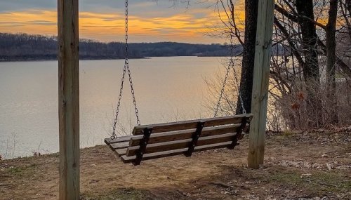 A wooden swing hangs from a frame by a calm lake at sunset, with trees lining the shore and a colorful sky.