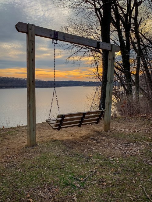 A wooden swing hangs from a frame by a lake at sunset, surrounded by trees and a tranquil landscape.