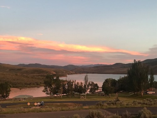 A serene landscape featuring a lake surrounded by hills and trees, with a colorful sunset in the sky.