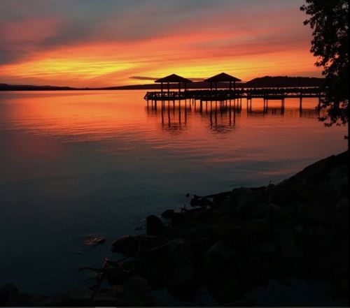 Silhouetted docks against a vibrant sunset reflecting on calm water, with a serene landscape in the background.