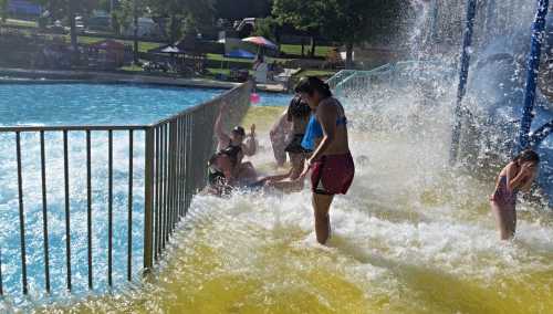 A water park scene with people enjoying a splash area, water cascading down, and colorful umbrellas in the background.