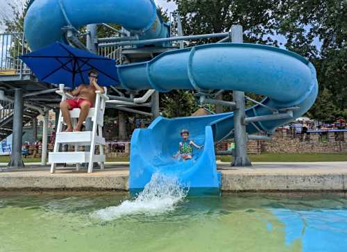 A child slides down a blue water slide while a lifeguard watches from a chair under an umbrella.