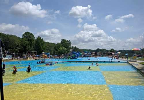 A vibrant outdoor pool area with people swimming, water slides, and colorful umbrellas under a partly cloudy sky.