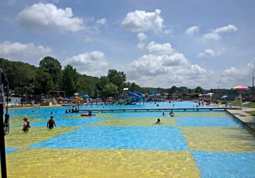 A vibrant outdoor pool area with people swimming, water slides, and colorful umbrellas under a partly cloudy sky.