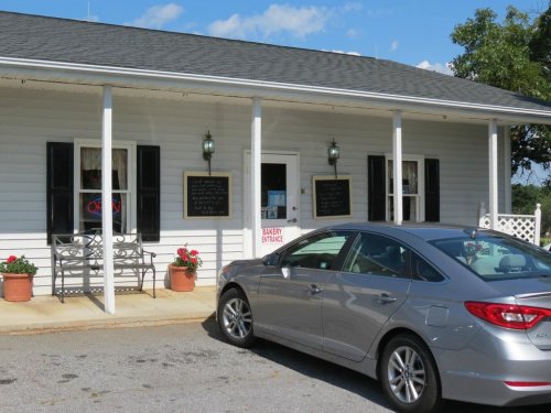 A silver car parked outside a bakery with a sign, flower pots, and a welcoming entrance on a sunny day.