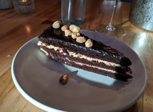A slice of chocolate cake with layers of cream, topped with crunchy nuts, on a white plate.
