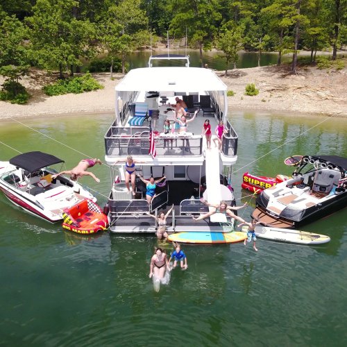 A group of people enjoying a sunny day on a houseboat, with boats and paddleboards nearby in a scenic lake setting.