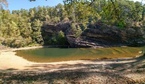 A serene riverbank scene with clear water, rocky cliffs, and lush greenery under a bright blue sky.