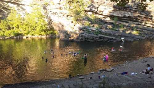 A group of people swimming and relaxing in a serene, rocky river surrounded by trees and sunlight.