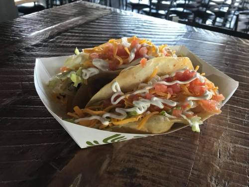 Three tacos filled with lettuce, tomatoes, cheese, and drizzled with sauce, served on a wooden table.