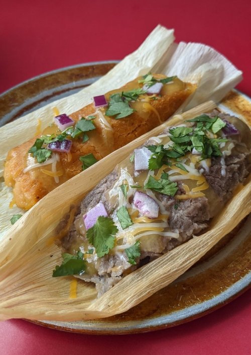 Two tamales on a plate, one with a golden crust and the other filled with meat, topped with cheese, cilantro, and diced onions.