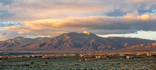 A panoramic view of mountains under a colorful sky, with a valley and scattered buildings in the foreground.