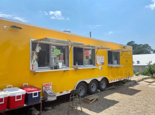A bright yellow food truck with open windows, parked in a gravel area, surrounded by coolers and outdoor seating.