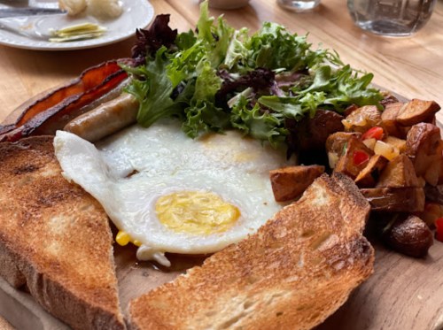 A hearty breakfast plate with fried egg, bacon, sausage, mixed greens, and seasoned potatoes on a wooden board.