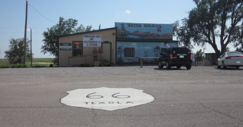 A roadside view of Water Hole 2, a store and restaurant, with a Route 66 sign in the foreground and trees nearby.