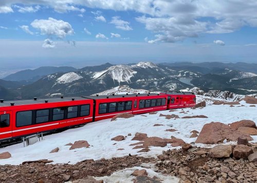 A red train on a snowy mountain landscape with distant peaks and a cloudy sky.