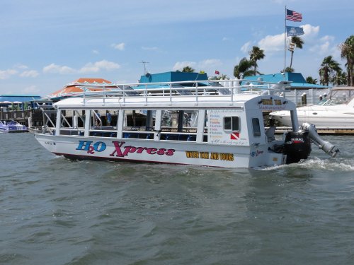 A tour boat named H2O Xpress navigates through water, with palm trees and buildings in the background.