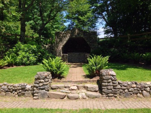 A stone structure surrounded by greenery and ferns, with a pathway leading to it in a serene garden setting.