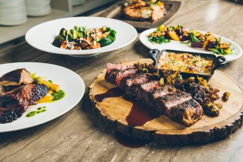 A wooden platter with grilled steak, surrounded by various gourmet dishes on white plates.