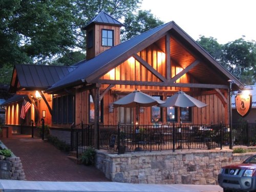 A cozy wooden restaurant with a stone facade, outdoor seating, and warm lights, set in a scenic area at dusk.