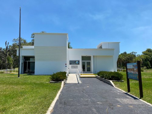 A modern white building with a pathway, surrounded by green grass and trees, featuring a sign in front.