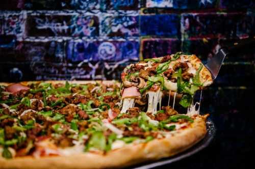 A slice of pizza topped with sausage, mushrooms, and greens being lifted from a large pizza against a colorful brick wall.