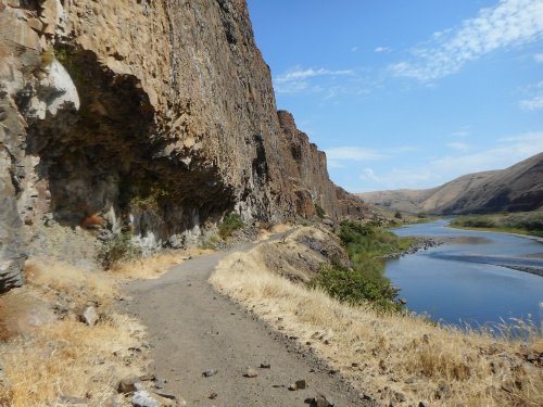 A winding dirt path along a river, flanked by steep rocky cliffs and dry grass under a clear blue sky.