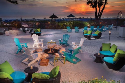 A beachside lounge area with colorful chairs and a fire pit, set against a vibrant sunset sky.