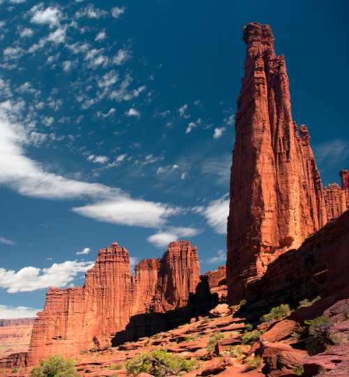 A striking red rock formation towers against a blue sky, surrounded by desert landscape and greenery.