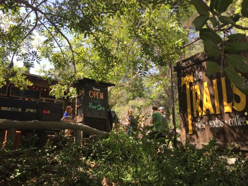 A rustic outdoor café sign surrounded by greenery, with people enjoying the space in a natural setting.