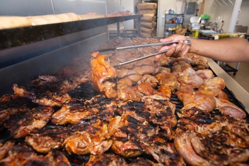 A hand using tongs lifts grilled meat from a large barbecue grill filled with various meats.