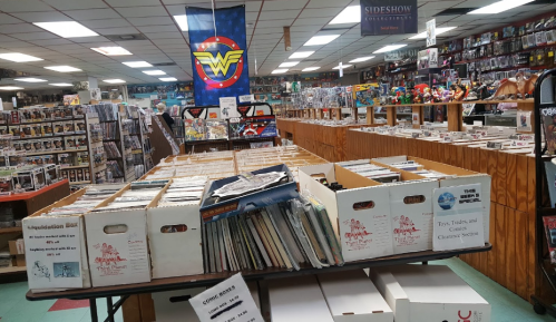 A comic book store interior with rows of boxes filled with comics and collectibles, featuring a Wonder Woman banner.