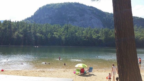 A serene lake scene with people swimming, a sandy beach, and a green umbrella under a mountain backdrop.