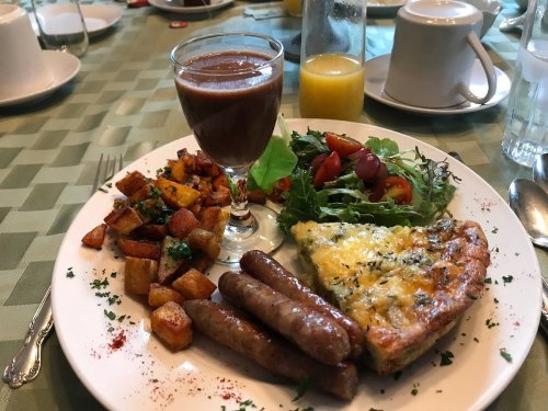 A plate with a frittata, sausages, roasted potatoes, a salad, and a glass of chocolate drink on a green tablecloth.