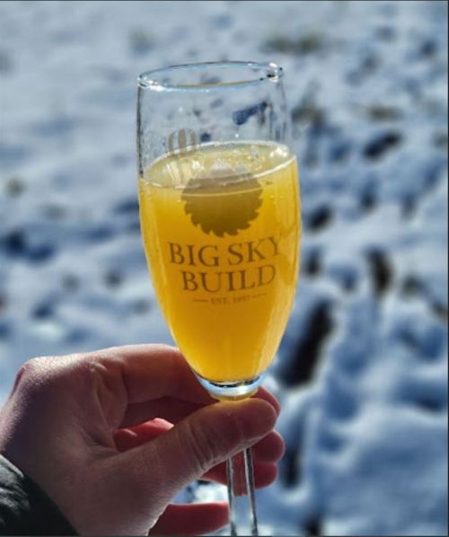 A hand holding a champagne flute filled with a yellow drink, with snow in the background. The glass has a logo.