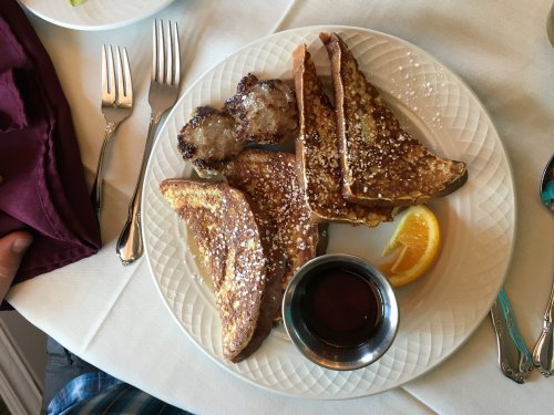 A plate of French toast with sausage, powdered sugar, an orange slice, and a small cup of syrup on a table setting.