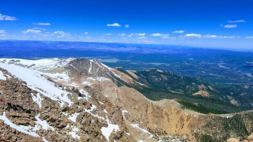 A panoramic view of rugged mountains and valleys under a clear blue sky with scattered clouds. Snow caps some peaks.