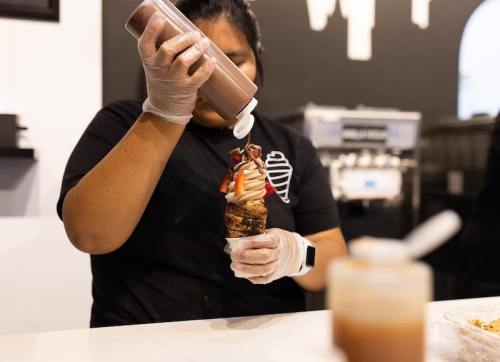A person in gloves drizzles chocolate sauce over a dessert cone topped with ice cream and strawberries.