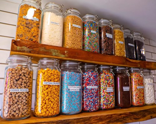 A shelf displaying various jars filled with colorful candies, cereals, and sprinkles, all labeled clearly.