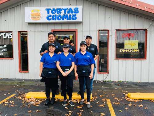 A group of five staff members stands outside Tortas Chomis, wearing blue shirts and black aprons, smiling at the camera.