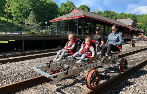 A family rides a pedal-powered rail cart on train tracks near a station surrounded by greenery.