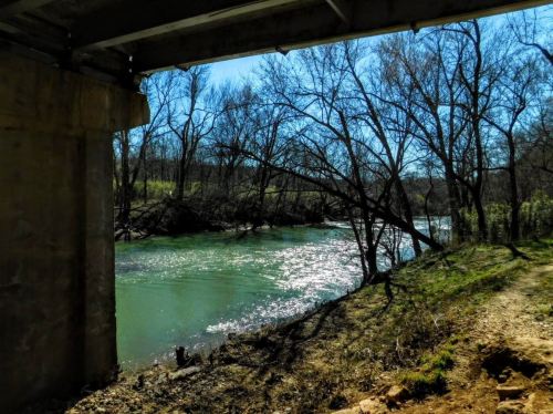 A serene river flows under a bridge, surrounded by trees and a clear blue sky. Sunlight sparkles on the water's surface.
