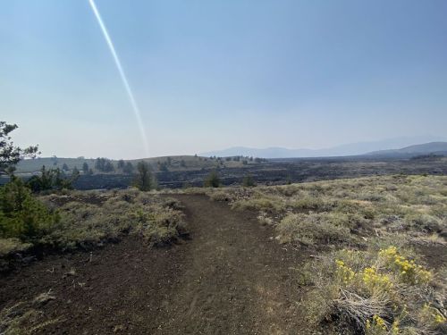 A dirt path winds through a barren landscape with sparse vegetation and distant hills under a hazy sky.
