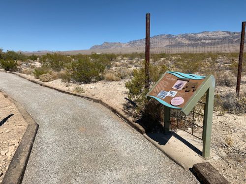 A gravel path leads to an informational sign in a desert landscape with mountains in the background.